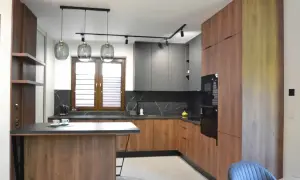 Nowoczesny, otwarty salon z jadalnią i kuchnią. Drewniane fronty szafek kuchennych i wyspa kontrastują z szarymi frontami górnych szafek.