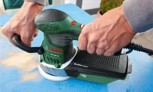 Dłonie trzymają zieloną szlifierkę Bosch PEX 400 AE, która jest szlifierką mimośrodową. Urządzenie pracuje na niebieskiej powierzchni.