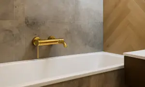 Elegancka, złota bateria zamontowana na środku wanny, nad jej krawędzią, w nowoczesnej łazience z szarymi i drewnianymi płytkami.
