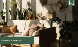 Przytulny salon w stylu boho z mnóstwem roślin, plecionym koszem i słomkowym kapeluszem na ścianie. Miękkie poduszki na kanapie zapraszają do relaksu.