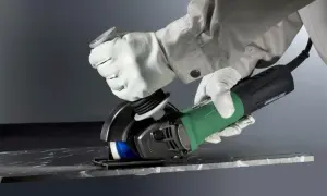 Ręce w białych rękawicach wykonują precyzyjne cięcie szlifierką kątową w ciemnym materiale.
