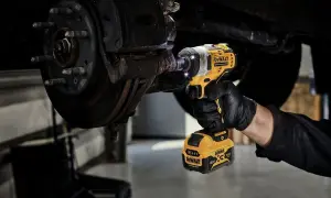 Ręka w czarnej rękawiczce używa żółto-czarnego klucza udarowego elektrycznego do odkręcania śrub przy kole samochodu.