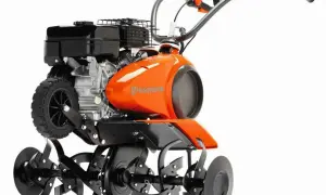 Pomarańczowy kultywator Husqvarna z silnikiem i obracającymi się nożami. Służy do spulchniania gleby w ogrodzie.