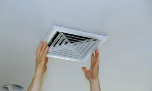 Montaż wentylacji łazienki w suficie. Dłonie instalatora przy białej kratce nawiewnej.