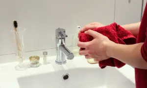 Domowe sposoby na czyszczenie płytek łazienkowych: ręce z czerwonym ręcznikiem przy białej umywalce, obok bateria i kosmetyki.