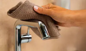 Dłoń z brązową ściereczką czyści chromowaną baterię łazienkową, pokazując, jak usunąć kamień z baterii łazienkowych.
