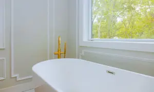 Biała wanna wolnostojąca, akrylowa czy stalowa, z elegancką złotą armaturą, obok okna z widokiem na drzewa.