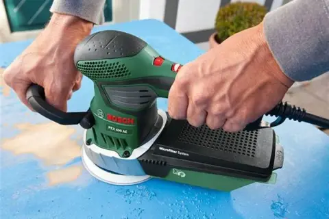 Dłonie trzymają zieloną szlifierkę Bosch PEX 400 AE, która jest szlifierką mimośrodową. Urządzenie pracuje na niebieskiej powierzchni.