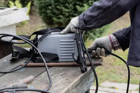 Ręce w rękawiczkach przygotowują spawarkę do domu. Widać uchwyt elektrody i kable.