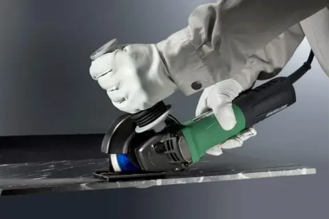 Ręce w białych rękawicach wykonują precyzyjne cięcie szlifierką kątową w ciemnym materiale.