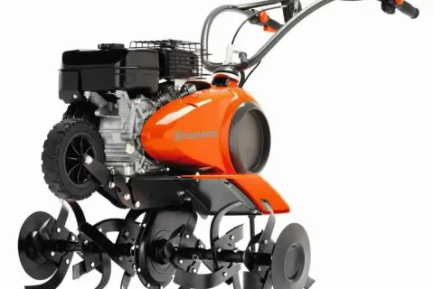 Pomarańczowy kultywator Husqvarna z silnikiem i obracającymi się nożami. Służy do spulchniania gleby w ogrodzie.