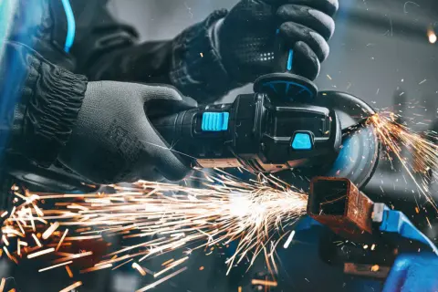 Ręce w rękawicach pracują przy budowie szlifierki kątowej, cięcie metalu generuje snop iskier.