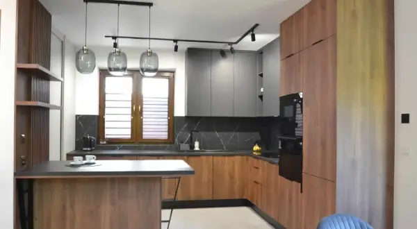 Nowoczesny, otwarty salon z jadalnią i kuchnią. Drewniane fronty szafek kuchennych i wyspa kontrastują z szarymi frontami górnych szafek.
