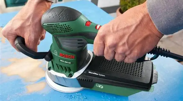 Dłonie trzymają zieloną szlifierkę Bosch PEX 400 AE, która jest szlifierką mimośrodową. Urządzenie pracuje na niebieskiej powierzchni.