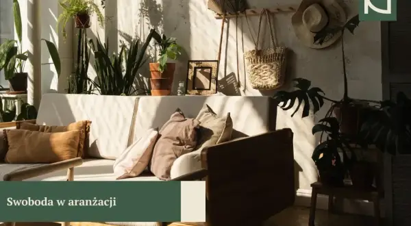Przytulny salon w stylu boho z mnóstwem roślin, plecionym koszem i słomkowym kapeluszem na ścianie. Miękkie poduszki na kanapie zapraszają do relaksu.