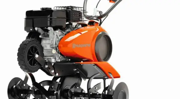 Pomarańczowy kultywator Husqvarna z silnikiem i obracającymi się nożami. Służy do spulchniania gleby w ogrodzie.