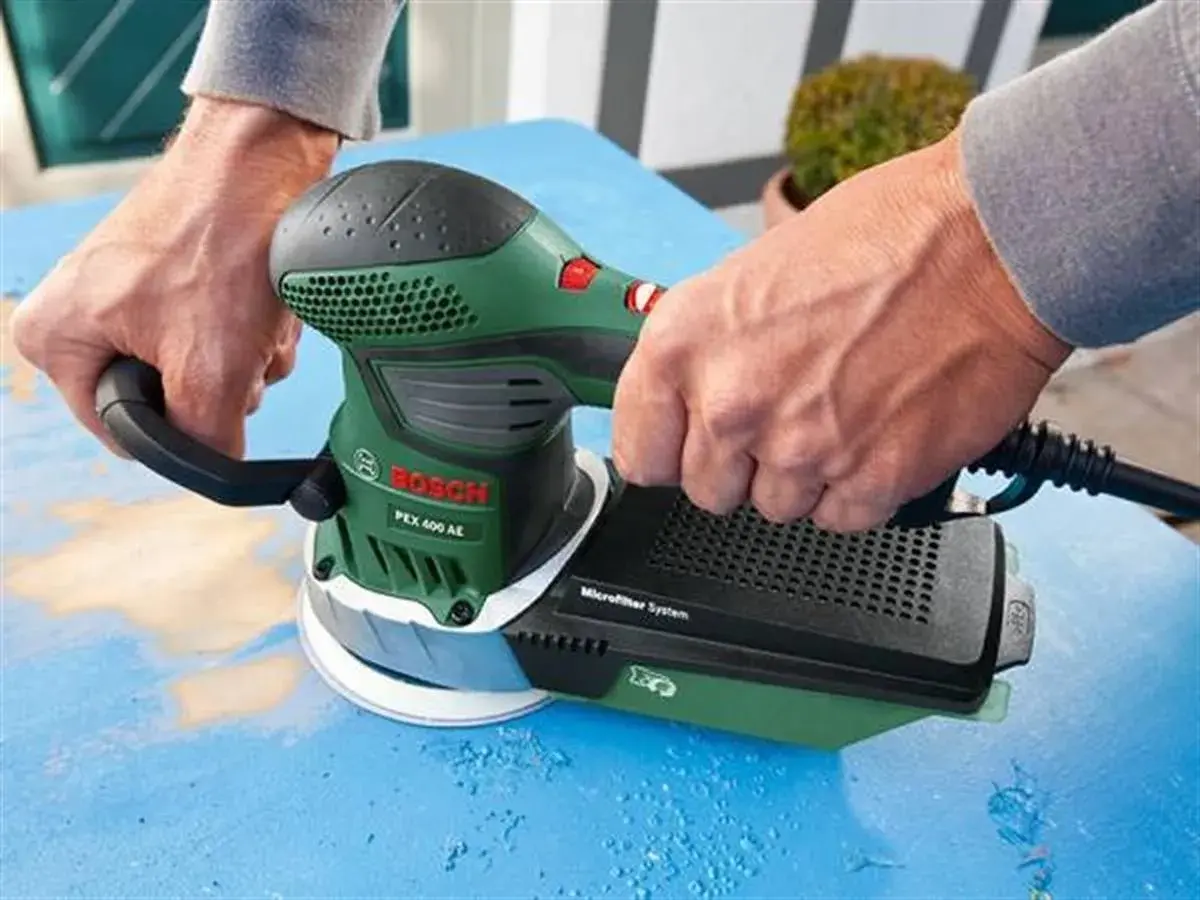 Dłonie trzymają zieloną szlifierkę Bosch PEX 400 AE, która jest szlifierką mimośrodową. Urządzenie pracuje na niebieskiej powierzchni.
