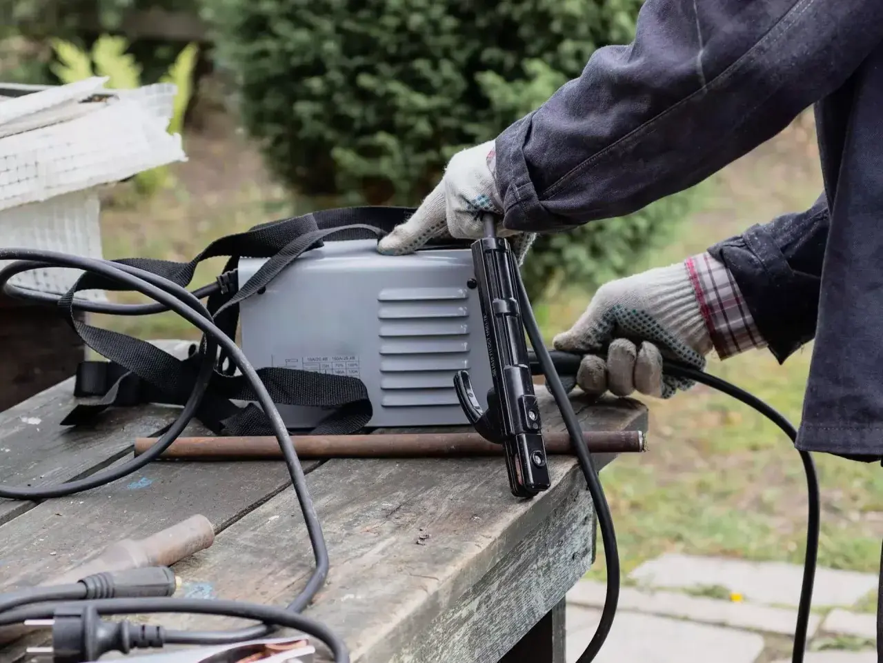 Ręce w rękawiczkach przygotowują spawarkę do domu. Widać uchwyt elektrody i kable.