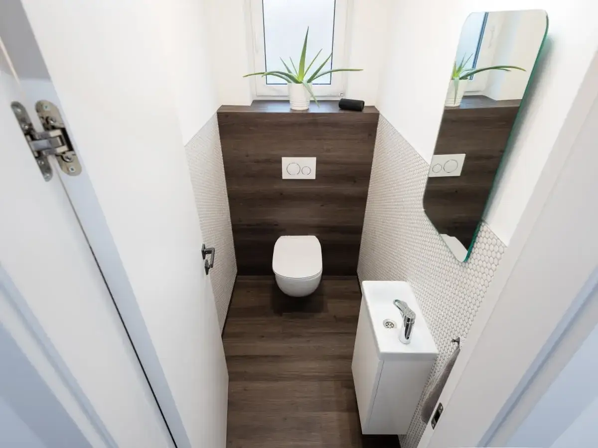 Nowoczesna łazienka z białym wc w rogu, małą umywalką i lustrem. Drewnopodobne płytki na podłodze i ścianie dodają ciepła.