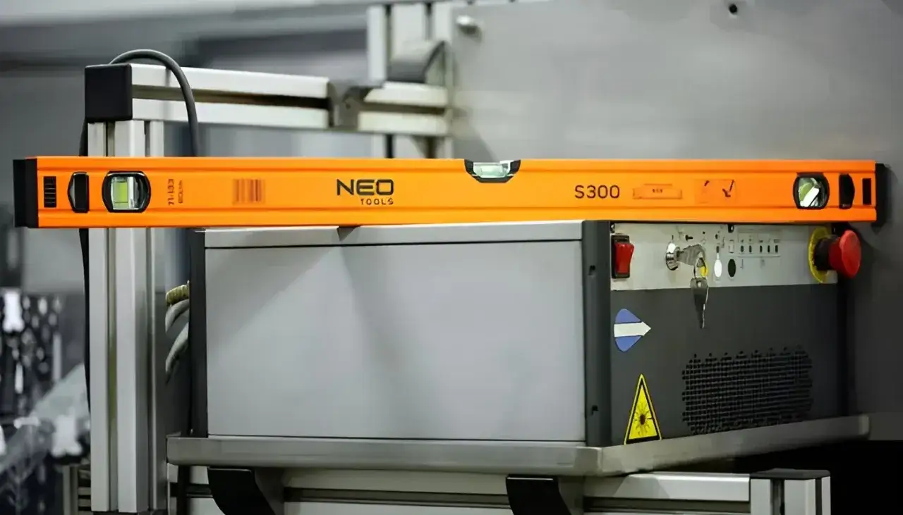 Pomarańczowa poziomica NEO TOOLS S300 leży na maszynie, sprawdzając jej idealne wypoziomowanie. Poziomica co to? Narzędzie do precyzyjnych pomiarów.