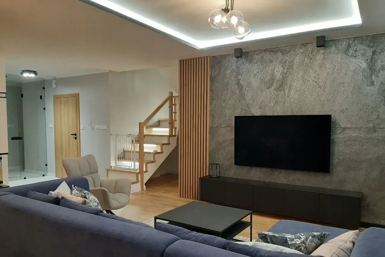 Nowoczesny salon z szarą kamienną ścianą, drewnianymi lamelami i wygodną kanapą. Świetne lamele ścienne inspiracje do Twojego wnętrza.