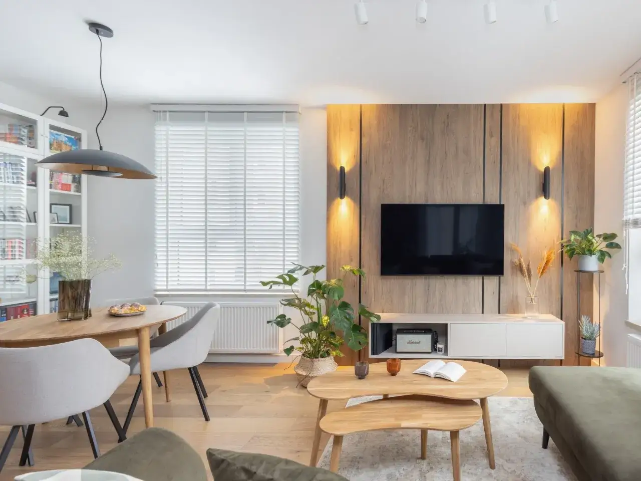 Przytulny salon inspiracje: drewniana ściana z TV, okrągły stół z szarymi krzesłami, rośliny i stylowe oświetlenie tworzą ciepłą atmosferę.