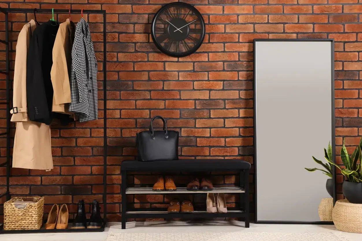 Przedpokój loft inspiracje: wieszak z płaszczami, ławka z butami, lustro i ceglana ściana tworzą stylową przestrzeń.