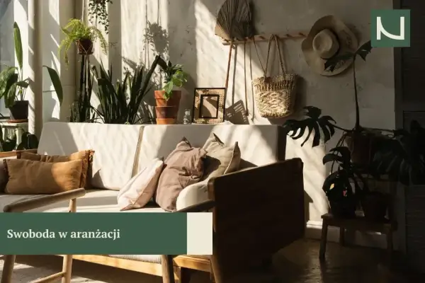 Przytulny salon w stylu boho z mnóstwem roślin, plecionym koszem i słomkowym kapeluszem na ścianie. Miękkie poduszki na kanapie zapraszają do relaksu.