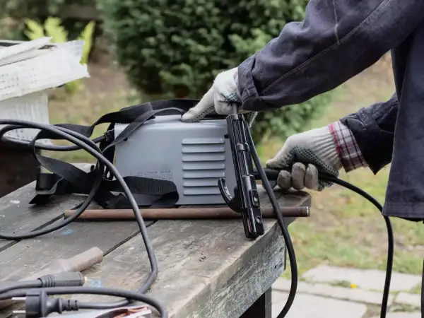 Ręce w rękawiczkach przygotowują spawarkę do domu. Widać uchwyt elektrody i kable.